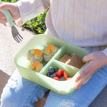 A child sits outdoors holding a green lunchbox with heart-shaped sandwiches, blueberries, strawberries, and a rectangular snack, wearing ripped jeans and a light sweater.