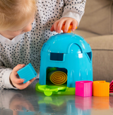 Child engaged with Fat Brain Toy Cos Shape Factory, honing spatial skills by fitting colorful blocks through openings on a reflective surface.