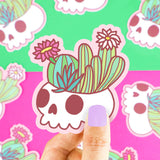 A hand holds a Turtle Soup Flowering Cactus Skull vinyl sticker, ideal for goth decor. Background is vibrant with plants.