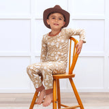 A boy in a cowboy hat smiles on a wooden chair in Tesa Babes Wild West Brown PJs, beige with white cactus patterns, eco-friendly.