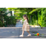 A young girl in a beige dress pulls the colorful PlanToys Pull Snail toy on a wooden pathway surrounded by greenery.