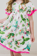 Girl in Mila and Rose’s Pom Pom Dress, Cute Cactus—ideal for a fun playground day with its floral print and pink pom-pom trim.
