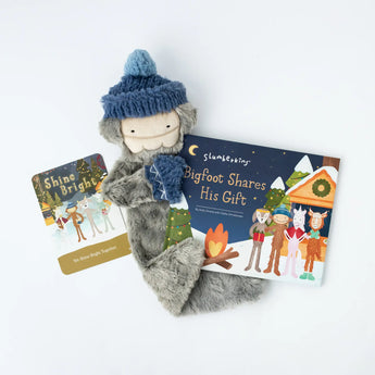 A Holiday Bigfoot Bundle Snuggler with a blue beanie, Bigfoot Shares His Gift book, and Shine Bright card from Slumberkins.