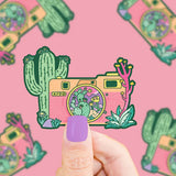 A person with lavender nail polish holds a Turtle Soup Vinyl Sticker, Desert Camera featuring a camera and cacti on a pink background.