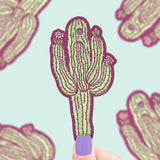 Purple nails hold a Turtle Soup Vinyl Sticker: Sonoran Saguaro against a light blue backdrop with cactus illustrations.