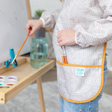 A child paints at an easel in a Bumkins Long-Sleeved Art Smock: Wander, with paints and water in a jar nearby.