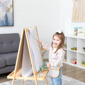 A girl with a bow in her hair paints at an easel, wearing a Bumkins Long-Sleeved Art Smock: Wander, in a cozy living room.