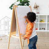 A child with curly hair draws on white easel paper in a bright room, wearing a Bumkins Long-Sleeved Art Smock: Grounded.