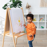 A child in a Bumkins Long-Sleeved Art Smock: Grounded paints at an easel, surrounded by shelves and plants in the bright room.