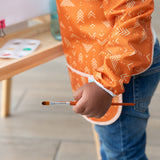 Close-up of a child wearing a Bumkins Long-Sleeved Art Smock: Grounded, holding a paintbrush next to an easel.