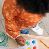 A child wearing a Bumkins Long-Sleeved Art Smock: Grounded uses a brush to paint with water and paints at an easel.