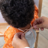 An adult fastens the neck tie of a Bumkins Long-Sleeved Art Smock: Grounded on a toddler.