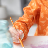 Close-up of a child in a Bumkins Long-Sleeved Art Smock: Grounded, painting beside a blurred glass jar.