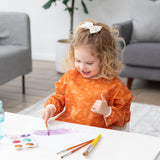 A smiling child with curly hair in a Bumkins Long-Sleeved Art Smock: Grounded paints on paper at a white table in a bright room.