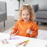A young child with curly hair paints watercolors at a white table while wearing a Bumkins Long-Sleeved Art Smock: Grounded.