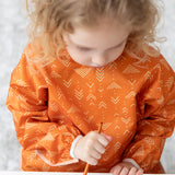 A child wears a Bumkins Long-Sleeved Art Smock: Grounded, paintbrush in hand, focused on painting.