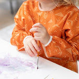 A child in a Bumkins Long-Sleeved Art Smock: Grounded paints with purple and gray paints on paper.