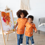Two children, one in a Bumkins Long-Sleeved Art Smock: Grounded, stand by an easel with decor including a pampas grass vase and gray chair.