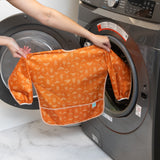 A person holds a Bumkins Long-Sleeved Art Smock: Grounded in front of an open front-loading washing machine.