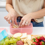 A person arranges crackers in a pink Bumkins Silicone Bento Box 3 Section: Pink Jelly with strawberries, grapes, and cheese on a board nearby.