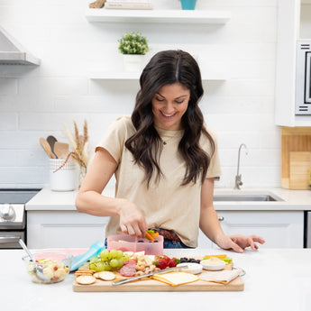 In the kitchen, a woman organizes snacks into a Bumkins Silicone Bento Box 3 Section: Pink Jelly on a fruit-lined counter.