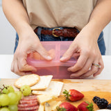 A person seals the lid of a Bumkins Silicone Bento Box 3 Section: Pink Jelly next to a cutting board with fruit, crackers, and cheese.