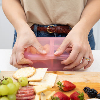 A person seals the lid of a Bumkins Silicone Bento Box 3 Section: Pink Jelly next to a cutting board with fruit, crackers, and cheese.