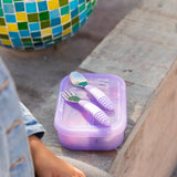 Bumkins Silicone Bento Box 3 Section: Purple Jelly with purple utensils sits on a bench next to a colorful mosaic pot.