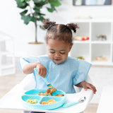 A young curly-haired girl sits in a high chair, wearing a Bumkins Ruffle Apron Bib: Cinderella while eating from a blue plate with utensils.
