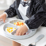A child enjoys eating with their hands from a divided plate while wearing a Bumkins Sleeved Bib: Jack Skellington.