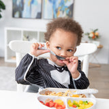 A toddler in Bumkins Sleeved Bib: Jack Skellington playfully holds a spoon to their mouth while sitting at a table with a plate of food.