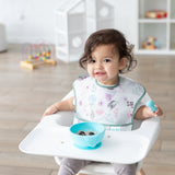 A toddler in a high chair wears a floral bib, eating oatmeal and berries from a Bumkins Silicone First Feeding Set: Blue in a bright room.