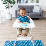 A baby eats from a Bumkins Silicone First Feeding Set: Blue, wearing a dark blue bib in a high chair with a matching mat on the floor.
