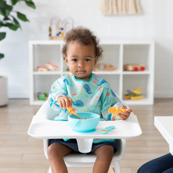 A toddler in a high chair in a Bumkins SuperBib® and Sleeved Bib 3-Piece Set: Rolling With The Waves, Whale Tail, Ocean eats from a blue bowl.