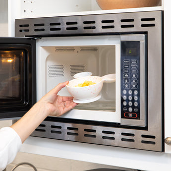 A hand places a Bumkins Silicone First Feeding Set: Mickey Mouse Vanilla Sprinkle filled with food into an open microwave.