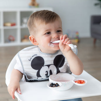 A toddler in a high chair enjoys yogurt and berries with a spoon from a Bumkins Silicone First Feeding Set: Mickey Mouse Vanilla Sprinkle.