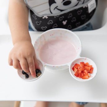 A child reaches for blueberries near yogurt and strawberries in a Bumkins Silicone First Feeding Set: Mickey Mouse Vanilla Sprinkle.