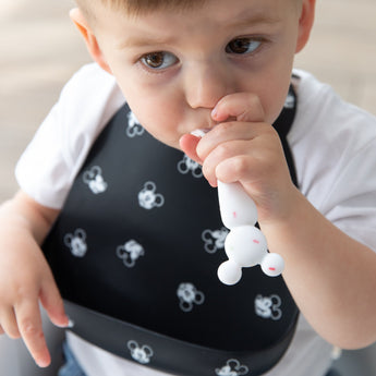 A toddler with brown hair wears a black bib while chewing a spoon from Bumkins Silicone First Feeding Set: Mickey Mouse Vanilla Sprinkle.