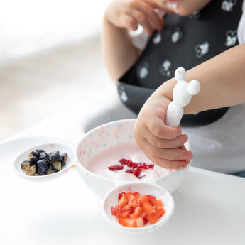 A child in a high chair enjoys yogurt with berries using a Bumkins Silicone First Feeding Set: Mickey Mouse Vanilla Sprinkle.