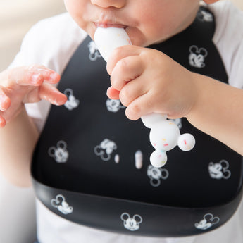A child chews a spoon from Bumkins Silicone First Feeding Set: Mickey Mouse Vanilla Sprinkle while wearing a black silicone bib.