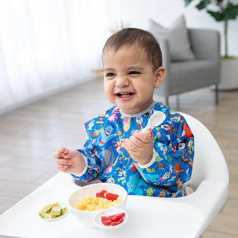 A toddler in a bib enjoys eggs, avocado and berries from a Bumkins Silicone First Feeding Set: Mickey Mouse Vanilla Sprinkle at a high chair.