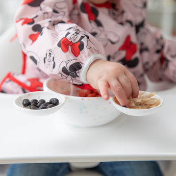 A child in a Minnie Mouse bib reaches for snacks from a Bumkins Silicone First Feeding Set: Mickey Mouse Vanilla Sprinkle on a high chair tray.