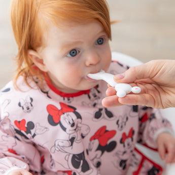 A red-haired toddler wearing a Minnie Mouse bib enjoys meal time with a Bumkins Silicone First Feeding Set: Mickey Mouse Vanilla Sprinkle.