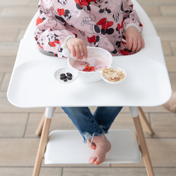 A child in a bib enjoys yogurt with berries from a Bumkins Silicone First Feeding Set: Mickey Mouse Vanilla Sprinkle on a high chair tray.