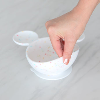 A hand lifts a Bumkins Silicone First Feeding Set: Mickey Mouse Vanilla Sprinkle on a marble surface, showcasing the suction cup bottom.