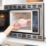 A person places a Bumkins Silicone First Feeding Set: Minnie Mouse Light Pink filled with yellow food into an open microwave.