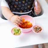 A toddler in a highchair eagerly explores the a Bumkins Silicone First Feeding Set: Minnie Mouse Light Pink, reaching for mashed sweet potato.
