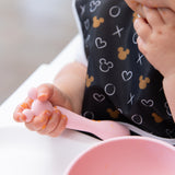 Close-up of a baby self-feeding with a Bumkins Silicone First Feeding Set: Minnie Mouse Light Pink, with food adorably smeared on tiny fingers.
