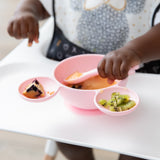 A child in a high chair enjoys using Bumkins Silicone First Feeding Set: Minnie Mouse Light Pink filled with kiwi, applesauce, and a muffin.