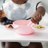 A baby sits in a high chair, enjoying mealtime with the Bumkins Silicone First Feeding Set: Minnie Mouse Light Pink.
